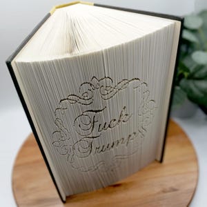 May include: A book with pages carved to display the words "Fuck Trump" within a decorative border. The book has a black spine and a gold-colored top edge. The book is displayed on a wooden surface.