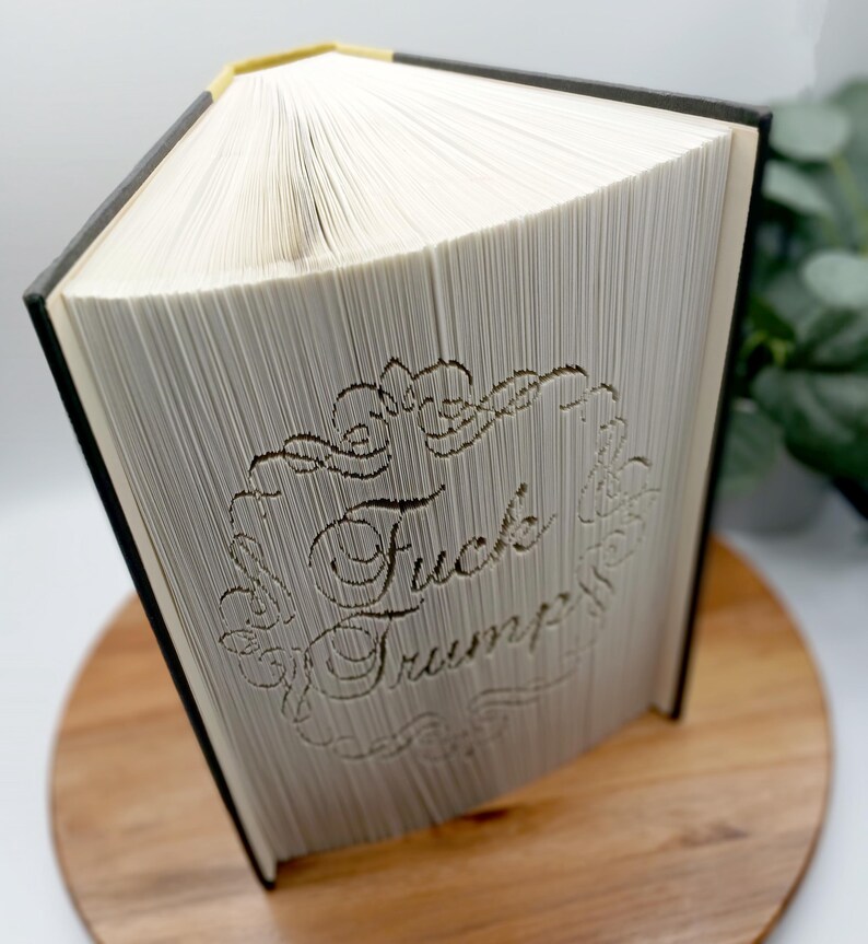 May include: A book with pages carved to display the words "Fuck Trump" within a decorative border. The book has a black spine and a gold-colored top edge. The book is displayed on a wooden surface.