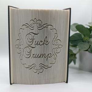 May include: A book with pages cut to reveal the words "Fuck Trump" within a decorative frame. The book has a black spine and the pages are a light beige color. The text is in a cursive font.