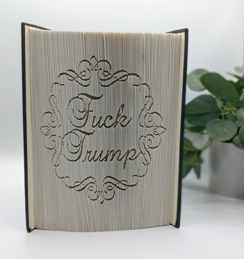 May include: A book with pages cut to reveal the words "Fuck Trump" within a decorative frame. The book has a black spine and the pages are a light beige color. The text is in a cursive font.
