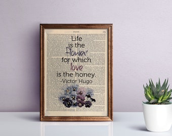 Puede incluir: Una impresión enmarcada sobre una página de diccionario antigua con la cita Life is the flower for which love is the honey de Victor Hugo. El texto está impreso en negro sobre un fondo de densas columnas de texto y flores pensamientos moradas.