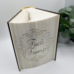 May include: A book with pages carved to display the words "Fuck Trump" within a decorative frame. The book has a black spine and gold-colored top edge. The pages are white, and the text is carved into the pages.
