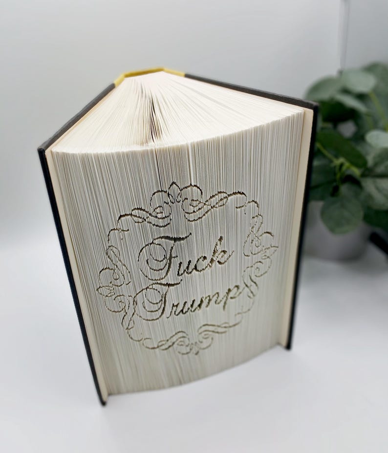 May include: A book with pages carved to display the words "Fuck Trump" within a decorative frame. The book has a black spine and gold-colored top edge. The pages are white, and the text is carved into the pages.