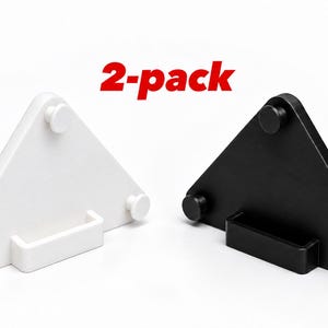 May include: A 2-pack of triangular wall-mounted shelves, one in white and one in black. Each shelf features a flat triangular surface, three small circular mounting pegs at the corners, and a rectangular ledge protruding from the bottom edge.