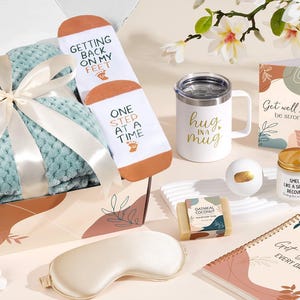 May include: A gift box containing a light green textured blanket with a white ribbon, white socks with text, a white mug with gold text, a greeting card, a crochet heart plant, a bath bomb, oatmeal soap, a candle, and a spiral notebook.