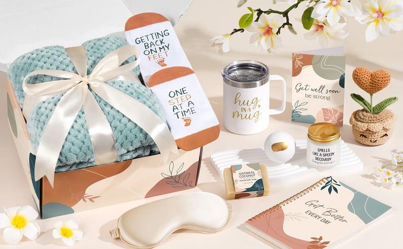 May include: A gift box containing a light green textured blanket with a white ribbon, white socks with text, a white mug with gold text, a greeting card, a crochet heart plant, a bath bomb, oatmeal soap, a candle, and a spiral notebook.