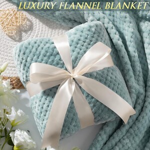 May include: A folded sage green flannel blanket with a textured waffle pattern, tied with a smooth white satin ribbon. The text LUXURY FLANNEL BLANKET is visible at the top. The blanket is soft and plush, featuring a consistent grid-like surface.