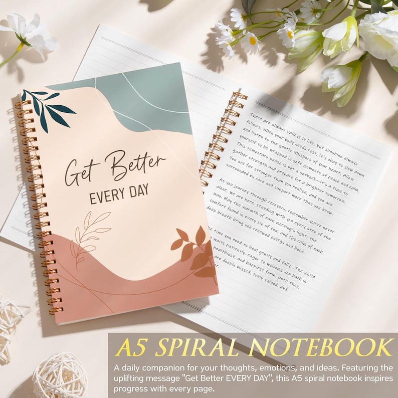May include: A spiral-bound notebook with a cover featuring abstract shapes in sage green, cream, and terracotta, with the text Get Better EVERY DAY. The open page displays printed text, and the notebook is accented with gold-toned metal spiral binding.