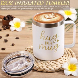 May include: A white 12oz insulated tumbler with a handle and a clear lid. The tumbler features the gold text "hug IN A mug" and is filled with coffee featuring latte art. The product is displayed with white plumeria flowers and coffee beans.