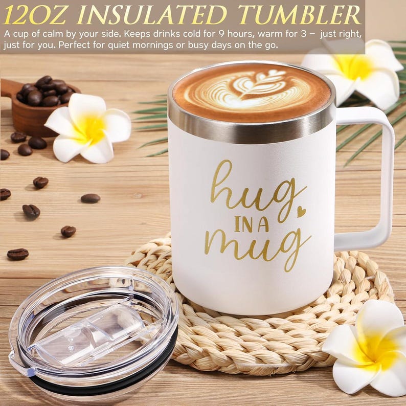 May include: A white 12oz insulated tumbler with a handle and a clear lid. The tumbler features the gold text "hug IN A mug" and is filled with coffee featuring latte art. The product is displayed with white plumeria flowers and coffee beans.