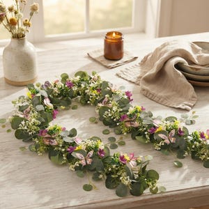 Puede incluir: Una guirnalda decorativa con hojas de eucalipto verdes, pequeñas flores blancas y moradas, y varios detalles de mariposas. La guirnalda está dispuesta en forma circular sobre una superficie de madera clara, con una vela y un jarrón cerca.