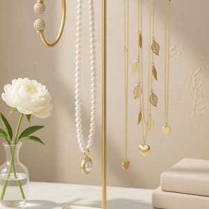 May include: A gold-toned jewelry display stand holding various necklaces, bracelets, and rings. The necklaces feature pearls and leaf or heart pendants. A white tray at the base holds rings and bracelets. A white flower in a vase is on the left.