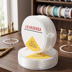 May include: Two stacked spools of clear elastic cord for jewelry making. The white labels feature the brand name LIS HEGENSA, a yellow triangle, and the text 0.7 and 70M/230ft. The spools are positioned on a wooden surface with jewelry supplies.