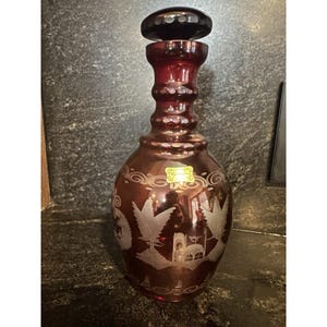 May include: A ruby red glass decanter with a black stopper. The decanter features etched designs of trees and buildings. The bottle has a bulbous base and a long, ridged neck. A small sticker is affixed to the front.