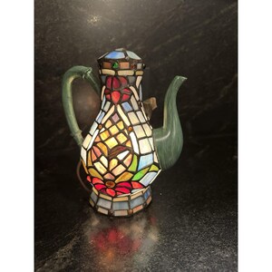 Könnte beinhalten: Eine dekorative Teekannenlampe mit einem Buntglasdesign. Die Lampe hat einen grünen Griff und Ausguss sowie ein farbenfrohes Mosaikmuster auf dem Körper. Das Buntglas umfasst rote, gelbe, blaue und grüne Farbtöne. Die Lampe ist beleuchtet.