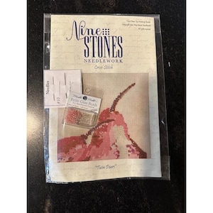 May include: A cross-stitch kit featuring a design of two pink pears. The kit includes needles, petite glass beads, and the text "Nine Stones Needlework Cross Stitch" and "Twin Pears". The kit is packaged in a clear plastic sleeve.