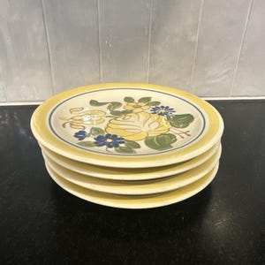 May include: A stack of four yellow ceramic plates. Each plate features a floral design with yellow roses, blue flowers, and green leaves. The plates have a yellow border with a thin blue line.