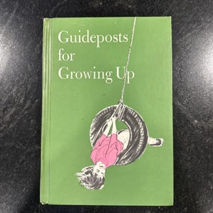 Może przedstawiać: Książka w twardej oprawie zatytułowana "Guideposts for Growing Up" z zieloną okładką. Ilustracja na okładce przedstawia dziewczynę w różowej koszuli i białych szortach huśtającą się na huśtawce z opony. Książka jest zamknięta.