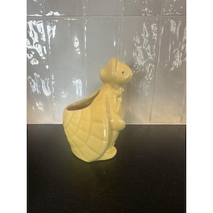 Può includere: Una fioriera a forma di coniglio in ceramica gialla con un design strutturato a forma di cestino. Il coniglio è in piedi con le orecchie alzate e una piccola apertura per piantare. La fioriera è su una superficie nera con uno sfondo piastrellato bianco.