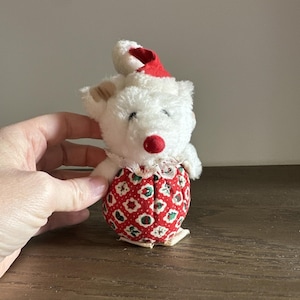 May include: A small, vintage stuffed bear with a white plush body, a red nose, and a red and white patterned outfit. It wears a red hat and has a white lace collar. The bear is a charming holiday decoration.