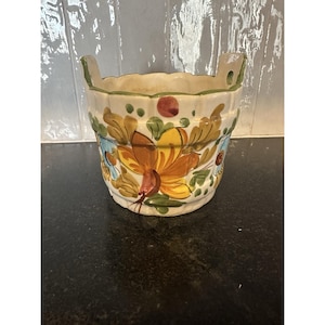 May include: A hand-painted ceramic planter with a cream-colored base and a floral design in shades of orange, yellow, blue, and green. The planter has two handles and a scalloped rim. The overall design is reminiscent of folk art.
