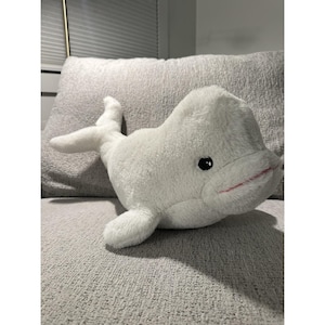 May include: A plush white beluga whale toy with black eyes and a pink mouth. The stuffed animal is lying on a gray textured surface, showcasing its soft, cuddly appearance. The toy has flippers and a tail.