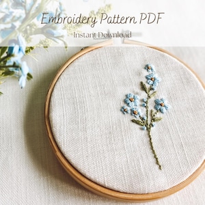 Peut inclure: Un cercle à broder en bois maintient un tissu blanc orné d'une broderie florale délicate. La broderie présente des fleurs bleu clair avec des centres jaunes et des tiges vertes. Le texte en haut indique "Embroidery Pattern PDF Instant Download."