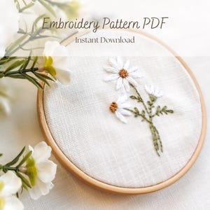 May include: An embroidery pattern PDF featuring a floral design of white daisies with brown centers and green stems. The design is set in a wooden embroidery hoop. Text on the image reads "Embroidery Pattern PDF" and "Instant Download."