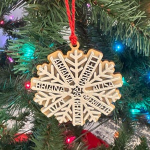May include: A snowflake-shaped wooden ornament featuring a layered design with a natural wood base and a white top layer. The ornament is engraved with the names Ariana, Barrett, Alisa, Merideth, Benson, and Briana, and hangs from a red string.