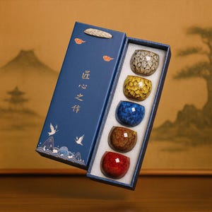 Peut inclure: Un ensemble de cinq tasses à thé en céramique colorées dans une boîte cadeau bleue. Les tasses sont dans des tons dorés, bleus, marron et rouges. La boîte présente des caractères chinois et des illustrations d'oiseaux et de montagnes.