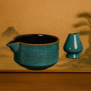May include: A teal ceramic tea bowl with a spout and a matching matcha whisk. The bowl has a dark interior and a textured exterior. The whisk is a small, rounded shape with a flat base. The set is displayed on a wooden surface.
