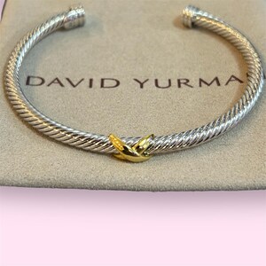 Könnte beinhalten: Ein silbernes Armband im Kabel-Stil mit gedrehtem Metalldesign und einem zentralen, polierten, goldfarbenen X-förmigen Akzent. Das Armband ist an den Enden offen und zeigt den Markennamen DAVID YURMAN in dunkler Schrift auf der Stoffoberfläche.