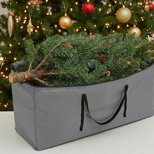 Könnte beinhalten: Eine graue Aufbewahrungstasche für einen Weihnachtsbaum mit schwarzen Griffen, die einen geschnittenen Baum enthält. Der Baum hat grüne Nadeln und kleine Tannenzapfen. Im Hintergrund ist ein dekorierter Weihnachtsbaum mit Lichtern und Ornamenten zu sehen.
