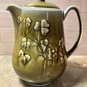 May include: Olive green ceramic pitcher with a lid and handle. The pitcher features raised white clover designs. The lid has a brown knob. The pitcher is on a light brown surface.