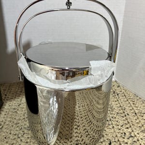 May include: A shiny, stainless steel ice bucket with a rounded body and a matching lid. It features a large, arched handle for carrying. The bucket is partially filled with white tissue paper, suggesting it is new and unused.