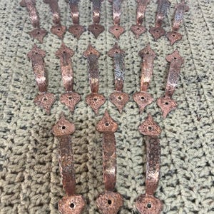 May include: A collection of copper-colored metal pulls with a hammered texture. Each pull has a decorative top and a teardrop-shaped base. The pulls are arranged on a textured, beige surface.