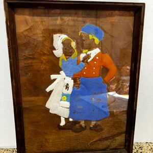 May include: Framed artwork featuring two figures in traditional attire. One figure wears a white bonnet and dress, the other an orange jacket and blue skirt. The artwork is set against a textured, brown background, and framed in a dark wooden frame.