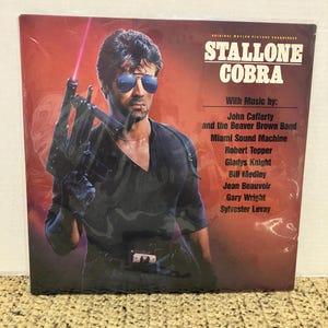 May include: A vinyl record album cover for the movie "Cobra" featuring Sylvester Stallone. The cover has a red background with Stallone in sunglasses holding a gun. The album title and music credits are also visible.