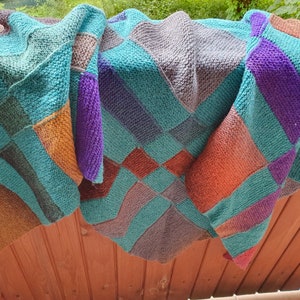 May include: A knitted blanket with a geometric design. The blanket displays squares and stripes in teal, purple, orange, brown, and grey. The blanket is draped over a wooden surface, highlighting its texture and colour variations.