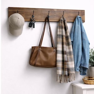 May include: A wooden wall-mounted coat rack with four black hooks. A tan baseball cap, keys, a brown leather tote bag, a plaid scarf, and a denim jacket hang from the hooks. The rack is mounted on a white wall.