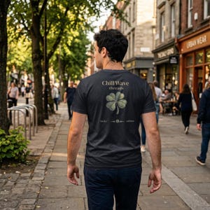 Può includere: Una maglietta grigio scuro con una grafica a quadrifoglio e la scritta "Chill Wave threads". La maglietta ha anche le parole "lucky" e "edition". La persona cammina su una strada lastricata.
