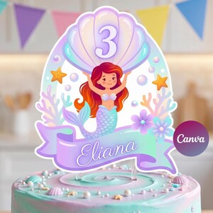 Op de afbeelding: Een cake topper met zeemeermint thema voor een derde verjaardag. Het ontwerp toont een cartoon zeemeermin met rood haar, die een schelp vasthoudt met het nummer 3. De naam "Eliana" staat op een banner. De topper staat op een pastelkleurige taart.