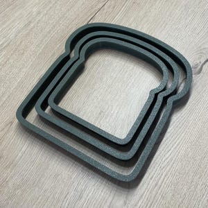 May include: Three nested, dark teal bread-shaped cookie cutters. The cutters are made of a textured material and are arranged on a light wood-grain surface. The cutters are designed to create bread-shaped cookies or sandwiches.