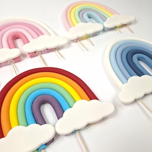 May include: Four rainbow-shaped cake toppers made of clay or fondant. One features bright primary colours, one has pastel shades, one is in shades of blue, and one is in shades of pink. Each rainbow has white clouds at the base and wooden sticks.