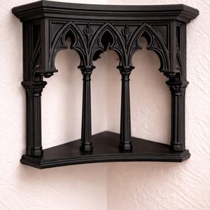 Könnte beinhalten: Ein mattschwarzes Eckregal mit einem dekorativen architektonischen Design, das vier vertikale Säulen und spitze, gotisch anmutende Bögen an der Oberkante aufweist. Das Regal hat eine dreieckige Basis und eine flache Oberfläche zur Montage.