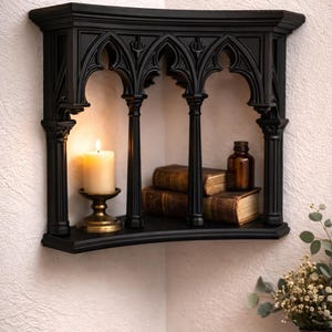 May include: A black corner shelf featuring an architectural design with pointed arches and vertical columns. The shelf holds a lit cream-colored pillar candle on a brass stand, two stacked antique books with worn covers, and a small brown glass bottle.