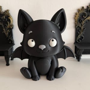 Peut inclure: Une figurine noire mate représentant une chauve-souris assise avec de grandes oreilles pointues, de grands yeux blancs tournés vers le haut et un petit nez gris. La chauve-souris a de petites ailes déployées et un ventre arrondi.