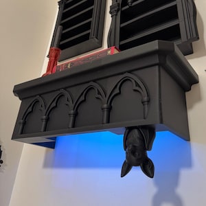 Könnte beinhalten: Ein schwarzes Wandregal mit gotischen Bogendetails an der Vorderkante. Eine skulptierte schwarze Fledermaus hängt kopfüber an der Unterseite des Regals, das von einem hellblauen LED-Lichtstreifen beleuchtet wird, der einen Schein nach unten wirft.