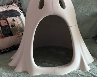 May include: A white, hollow, dome-shaped structure resembling a ghost with two oval-shaped eye cutouts and a large, arched opening at the base. The object has a smooth, matte finish and a curved, flowing bottom edge that mimics the appearance of a sheet.