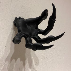 Könnte beinhalten: Eine schwarze, stilisierte Handskulptur mit langen, gebogenen Krallen, die an einer strukturierten weißen Wand befestigt ist. Der Schatten der Hand ist sichtbar. Das Design deutet auf ein Fantasy- oder Gothic-Thema hin.